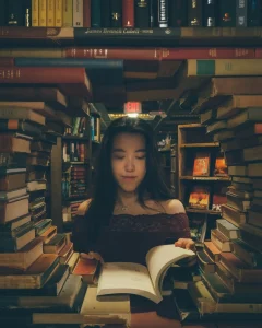 Photo of a young woman with straight black hair and olive skin standing in a bookstore. Her ethnicity isn’t perfectly clear, but she could be Asian or Middle Eastern. She’s reading a hardback novel while standing in front of a display of books that has been arranged in a large circle formation that perfectly frames her chest, shoulders, and head. It gives the effect of looking through a mirror or a portal and seeing her on the other side. It’s very cool.