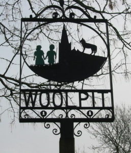 An iron sign that has the name Wool Pit carved out of iron in it. The sign also has a carving of two children, a church, and a wolf on it. The rest of the sign is comprised of thin rods that are needed to hold everything together. You can see a great deal of the grey sky and the empty tree branches through it. 