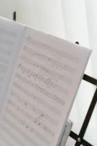 A close-up of a sheet of music on a music stand. The stand is next to a window that is covered by horizontal blinds. 