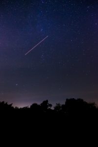 A shooting star zipping across a velvety purple and black night sky just after sunset. You can see the stars beginning to twinkle in the sky and the dim outline of a forest in the background where the last drops of sunlight are quickly fading out of view. 