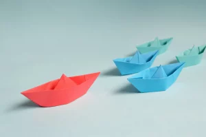 Five little paper boats sitting on a light blue surface. The one out in front is red, the two behind that one are blue, and the last two are teal. 