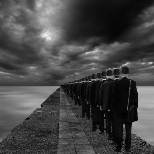 Surreal photo of dozens of people wearing suits and the same bowl-shaped hats standing in a neat row on a sidewalk under a stormy sky and next to such a thick layer of fog you can’t tell if they’re many miles up into the air on this surface or if there’s a calm little sea just below the fog. 