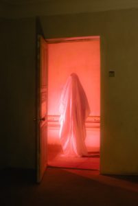 a hazy pink photo of a ghost standing in a hallway that’s lit by a pink light. The viewer is looking at it from the perspective of a pitch black room where nothing else can be seen in the foreground. 