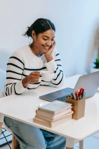 A brown woman sitting at a desk and grinning while holding up her head with her left hand. She’s looking at something on her laptop. She is wearing a white and black striped sweater and holding a white mug in front of her with her left hand. 