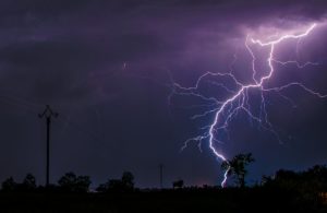 A dramatic photo of lighting striking the ground just after sunset. You can see the dim outline of a tree and a house on the land, but everything else is dark. The sky above is partially black, partially purple near where the lightning is striking, and partially lit up from the lighting itself. 