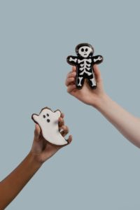 Two white people are each holding up a sugar cookie that has been decorated for Halloween. One cookie is shaped like a ghost and the other one a skeleton. 