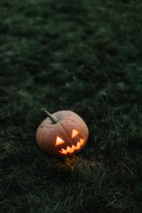 A jack-o-lantern is sitting in a patch of glass. There is a candle inside of the lantern that is making it glow orange and yellow. It’s dark outside, so few other details can be seen other than the lush grass it is sitting on. 
