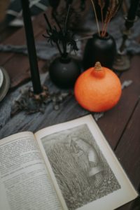 An opened book is sitting next to a small pumpkin on a dusty wooden table. There is an illustration of some sort of gnome or other heavily-bearded figure on the right hand side of the page. It looks like an illustration from a dark fairy tale, maybe? Behind the book and pumpkin is a black candle and some orange and black sticks sitting in black pots. 