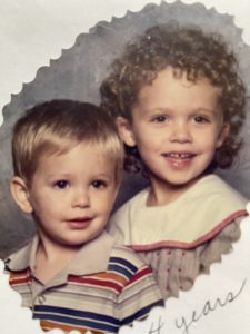 A 1980s photo of two young siblings, ages about 4 and 2, who have been posed for a professional photo. The older girl child is me, Lydia. She has short, very curly brown hair and is wearing a light purple dress that has a large white collar with red trim on it. The little boy is my brother. He has short, straight, blond hair and is wearing a collared shirt that has a rainbow pattern. 