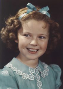 A photo of Shirley Temple when she was a little girl, maybe 6 years old. She’s grinning and wearing a blue dress with white lace trimming the collar and front portion of the dress. She has a cute little blue bow tied up around her curls, too. 