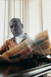 A black man sitting at a table and reading a newspaper. He has a serious, thoughtful expression on his face and has just looked up to make eye contact with the reader when this shot was taken. He’s sitting bedside a large picture window that has the blinds drawn, but it’s such a sunny day that you can still see lots of light pouring into the room. 