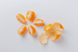 A peeled mandarin orange has been broken into five segments. It’s sitting on a white surface next to the mandarin orange peel that once held this fruit. 