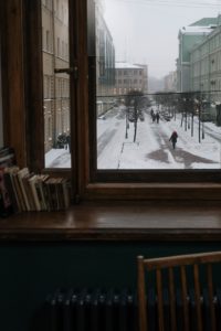 Hardback books neatly lined up on a wooden desk that is sitting right in front of a large picture window. The window overlooks a snowy winter street where a pedestrian is walking down the centre of the road because the sidewalks are filled with snow. 