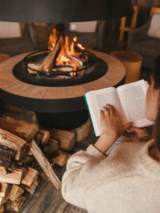 A white woman who has brown hair and is wearing a chunky white sweater is reading a hardback novel as she sits next to a fireplace. The fireplace is in a circular fire pit in the centre of the room. You can see a stack of chopped wood next to it ready to add to the flame as needed. 