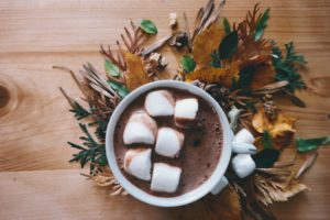 A photo of a white cup filled with hot chocolate and six big marshmallows. the cup is sitting on a wreath of pine branches and brown autumn leaves that have been arranged around it in a circular pattern. There are three extra marshmallows on this wreath, and everything is sitting on a wooden table. 