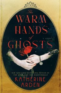 Book cover for The Warm Hands of Ghosts  by Katherine Arden. Image on cover shows a pair of painfully pale hands clasping a red rose. They have a bracelet with a small pendant on their left wrist, and both hands are partially covered by the leaves and branches that are concealing the rest of this person’s body. 