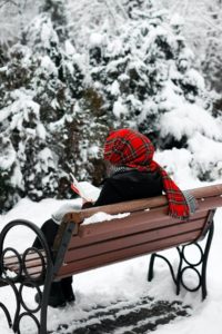 A person who is all wrapped up in a a warm winter coat and a red and black headscarf is sitting on a snowy bench in a park reading a book. You can see evergreen trees covered in a thick layer of snow in front of them, too. 