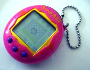 A photo of a Tamagotchi toy that was popular in the late 1990s and early 2000s. It’s made of pink plastic and has a tiny little screen where a pixelated pet can be seen sitting in the centre of the screen. 
