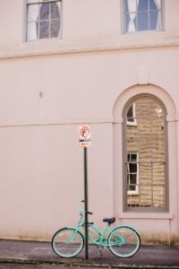 A teal bicycle that’s Ben chained to a no-parking sign on a quiet city street. The building behind the bicycle has been painted a beautiful shade of light pink. 