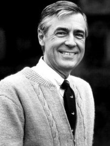 A black and white photo of Fred Rogers that looks like it was taken in the 1960s or 1970s. He is smiling ,has a full head of hair, and is wearing a white shirt and tie underneath a cabled sweater. 