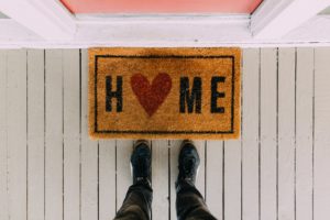 A photo looking down at someone’s legs as they stand on a porch next to a welcome mat that has the word “home” written on it in a thick black font. The “o” in the letter home has been replaced with a red heart. 