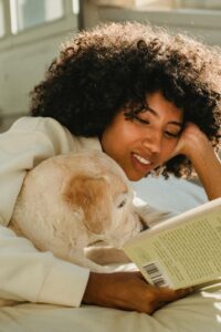 A brown-skinned woman with gorgeous, curly, black hair is cuddling her yellow Labrador retriever as she reads a book. The dog looks interested in the book. 