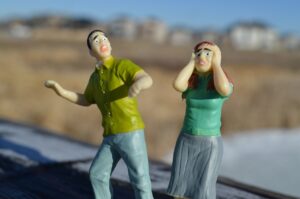 A photo of two little white figurines wearing jeans and two different shades of green shirts who look comically scared. Their facial features and body language are seriously over-exaggerated. 