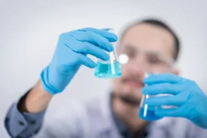 An out-of-focus photo of a young Hispanic doctor or researcher swirling a blue liquid around in one beaker while his other hand holds another beaker filled with blue liquid still. 