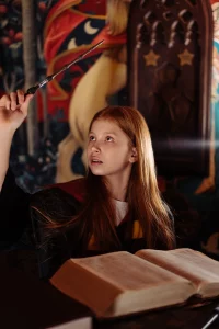 A young red headed girl is reading a book and attempting to cast a spell with a wooden wand. She’s holding the wand above her head and looking expectedly for some sign it’s working! 