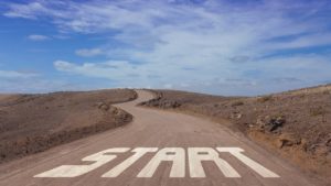 A photo of the word “start” painted in large white letters on an otherwise empty country road. The road is brown and dusty. The grass beside it on both sides is dead. The sky above is blue and partially cloudy. 