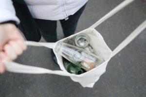 Close-up photo of a white person holding open a white canvas bag. Inside the bag are an assortment of glass and aluminum bottles. 