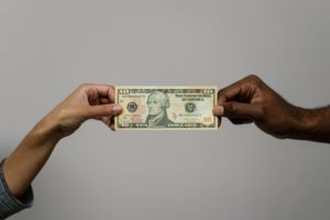 A photo of the hands of two people who are holding one side each of a $20 US bill. One of the people has dark skin and the other person has a medium skin tone. 