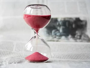 Pink sand moving through an hourglass that’s sitting on a white surface in front of a white wall that has a black abstract painting hung upon it. 