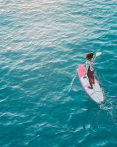 Aerial shot of a black woman paddle boarding on a pink and white paddle board in a green-blue sea. This means that she is standing up on a surfboard-like item and holding a paddle as she looks straight ahead. The water is rippling slightly in the wind and looks gorgeous. The woman has a confident pose and seems to be having a great time. 