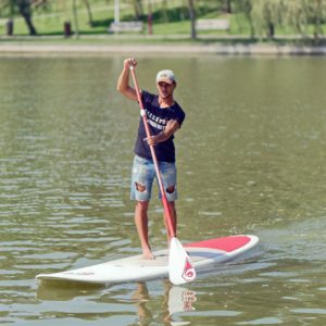 A white man paddle boarding in a green, algae-filled lake. He is wearing a t-shirt, a pair of jean shorts that has large holes in them, and a white baseball cap. His paddle board has a patch of reddish-orange colour on its otherwise white colour. The man looks like he’s having a good time but that he’s also carefully looking at something just out of view. His posture is slightly slouched to the left as he stands on his board. I do not know if that is significant or not, but thought I’d better include it in case it is. 