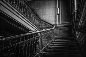 A black-and-white photo of a cobweb-covered staircase in an old house. On vthe right you see the stairs up to the landing. To the left you see the railing for the next flight of stairs up to the next floor, but you cannot see the stairs themselves due to how the staircase curves after the landing. 