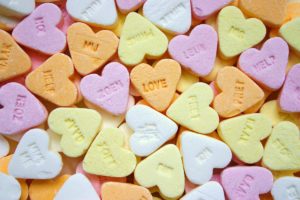 Closeup photo of pink, yellow, orange, and white conversation hearts. Not all of their messages can be read, but the ones that can be say things like “love,” “soft,” and “help.” 