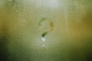A question mark on a foggy window. It looks like it was made by someone’s finger recently. You can see something green and fuzzy through the foggy window that might be a forest, but it’s too obscured to know for sure. 