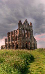 A photo of an abandoned stone cathedral on a grassy hill. It still has walls but no longer has a roof or glass in its windows. There is a stormy grey and slightly pink sky behind it. 