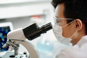 A male Asian scientist wearing a face mask and peering through a microscope at something on a slide. Perhaps he is looking at a highly infectious disease?