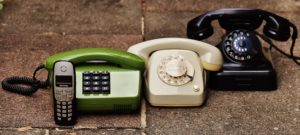 A photo of four different phones. From left to right there is a: 2000s-era black cellphone, 1980s-era green landline, 1960s-era white rotary phone, and 1940s-era black rotary phone. 