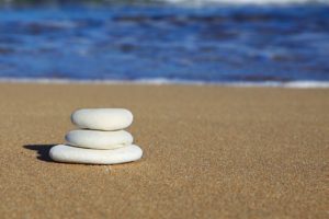 Three flat, white stones stacked on top of each other on the perfectly smooth sand of a beach. You can see the lake or ocean water in the distance. 