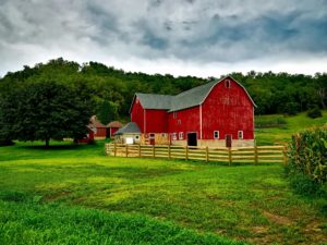 Photo of a red barn and a red farmhouse. There is a grassy field in the foreground and a nice, big forest behind the house. The sky overhead is partly cloudy. 