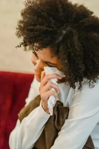 Photo of a biracial woman dabbing away tears from her cheek as she cries. She is wearing a white, longsleeved blouse and has a lovely Afro. 
