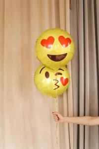A photo of the arm of a pale-skinned person who is holding two balloons against some beige drapes. Both balloons are yellow. One shows a kissing face emoji and the other shows the emoji whose eyes have been replaced with two little red hearts. What a cute scene!