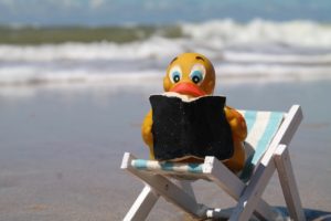 A adorable little figurine of a yellow rubber ducky who is sitting in a beach chair and reading a book. This figurine has been placed on a beach, and you can see a wave gently reaching the shore in the distance on this calm and sunny day. 