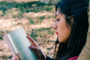 Closeup photo of a pale-skinned woman with long, black, straight hair reading a book. She’s sitting outdoors next to grass that is partially green and partially brown and dry. It looks like grass that has gone dormant in the summer heat!