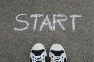 A photo looking down at a pair of black and white running shoes. The person’s feet (and shoes) are standing on a grey surface where the word “start” has been written in chalk. 