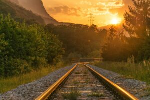 Photo of train tracks going through a forest. There isn’t currently a train on them, but you can see the sun setting in the distance and the dark, evergreen forest on either side of the tracks. It’s a quiet and thoughtful moment. 