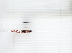 Stock photo of a white person peeking out at the world through white blinds. 